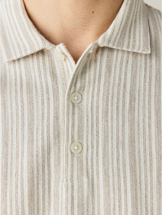 Close-up van een beige polo met verticale, fijne strepen in beige en gebroken wit. Twee knopen zichtbaar.