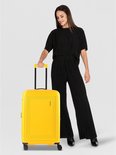 Vrouw met zwarte outfit staat naast gele American Tourister koffer.