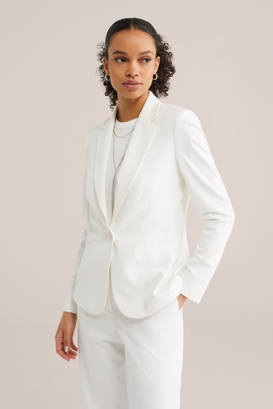 Model draagt een witte getailleerde blazer met een zilverkleurige ketting en een witte top.