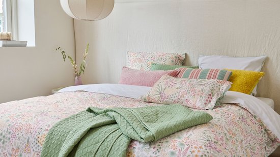 Een opgemaakt bed met een dekbedovertrek met botanische print, een groene sprei, en diverse sierkussens in pastelkleuren en met streepjes.