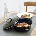 Een blauw-zwarte braadpan met een deksel ernaast, gevuld met vlees en aardappelen. Een fles water en een stoel staan op de achtergrond.