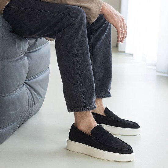 Un homme assis porte un haut beige, un jean gris et des mocassins noirs en daim à semelles blanches.