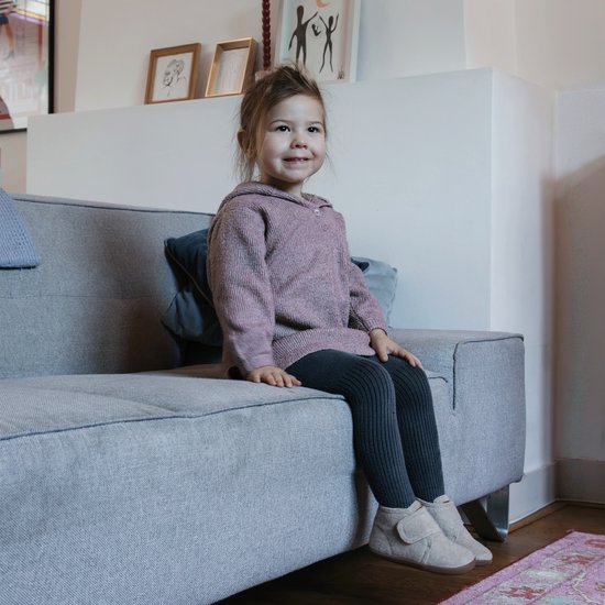 Een jong meisje zit op een grijze bank, gekleed in een roze trui en grijze legging.