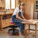 Vrouw in spijkerbroek en schort zit op rode rolkruk, werkend aan een houten plank in een werkplaats.
