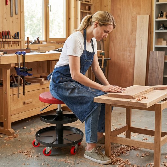 Vrouw in spijkerbroek en schort zit op rode rolkruk, werkend aan een houten plank in een werkplaats.