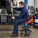 Monteur in blauw werkpak zit op een rode rolkruk met gereedschap onder een auto.