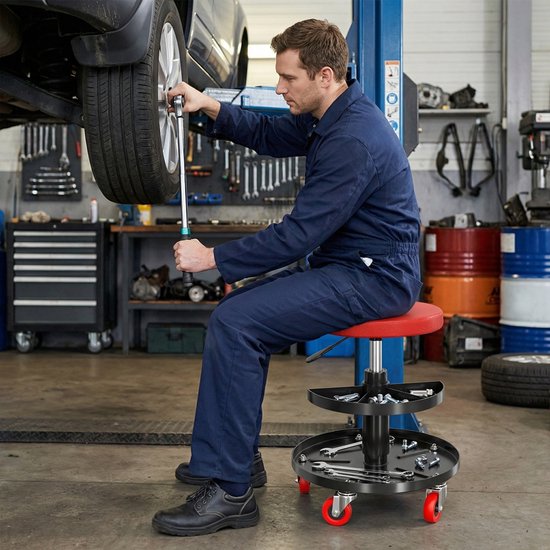 Monteur in blauw werkpak zit op een rode rolkruk met gereedschap onder een auto.