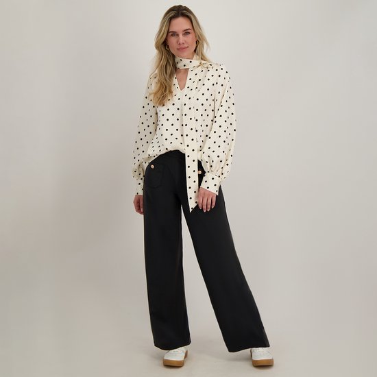 Vrouw in zwarte wijde broek en crèmekleurige blouse met zwarte stippen en strik.