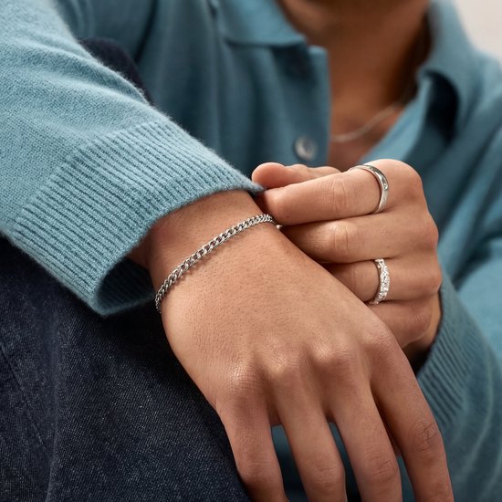Gros plan d'un bras portant un bracelet chaîne gourmette argenté et deux bagues. Pull gris.