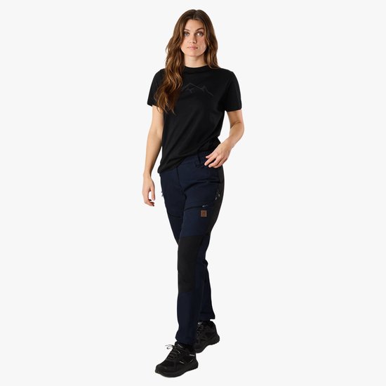 Vrouw in donkerblauwe outdoorbroek met zwarte panelen en een zwart T-shirt met bergprint.