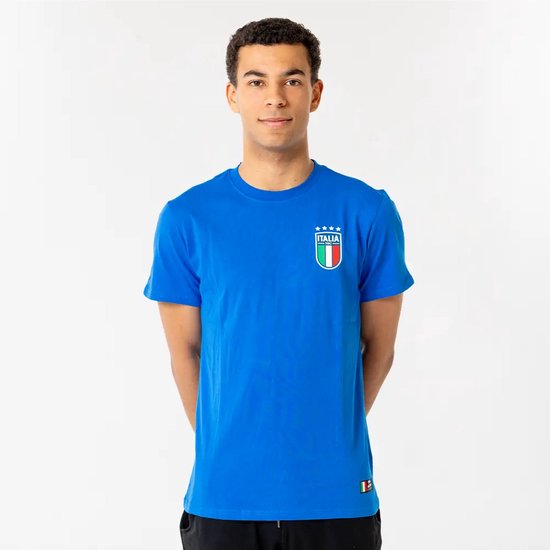 Un jeune homme porte un t-shirt bleu avec le logo 'Italia', quatre étoiles et le drapeau italien sur la poitrine. Les mains jointes derrière le dos.