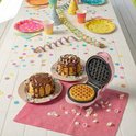 Roze mini wafelijzer op een feestelijk gedekte tafel met pannenkoeken en confettie.