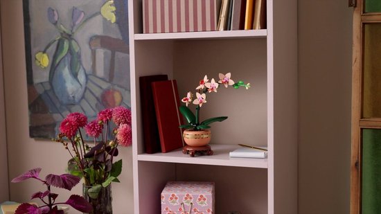 LEGO orchidee in een pot op een boekenplank, naast boeken en een vaas met bloemen.