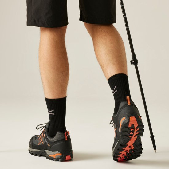 Man met zwarte wandelschoenen en oranje accenten, staand met wandelstok.