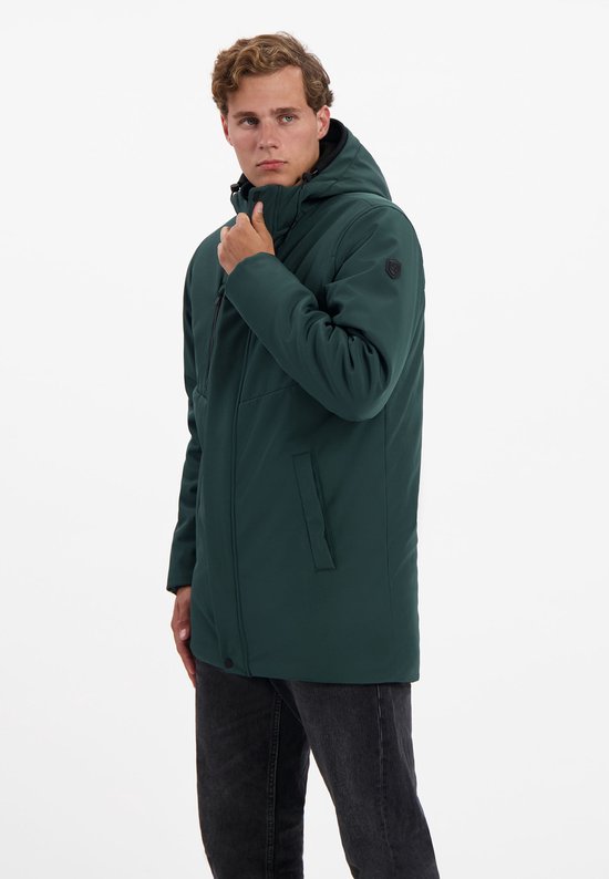 Man met donkergroene outdoorjas met capuchon, hand op de kin, zwarte spijkerbroek.