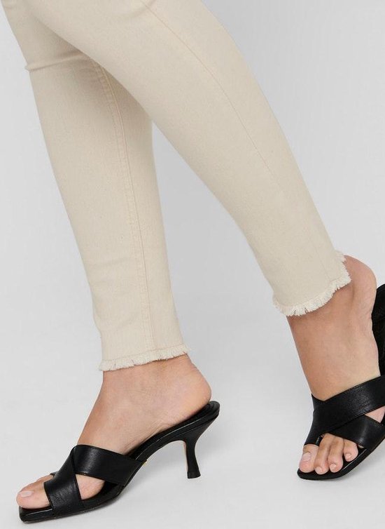 Close-up van beige skinny jeans met rafelige zoom en zwarte muiltjes met bandjes.