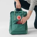 Groene Fjällräven Kånken rugzak met een hand die paspoorten in een voorvak steekt.