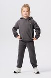 Meisje in donkergrijze hoodie met logopatch en joggingbroek. Handen in de zij. Witte sneakers.