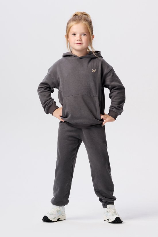 Meisje in donkergrijze hoodie met logopatch en joggingbroek. Handen in de zij. Witte sneakers.