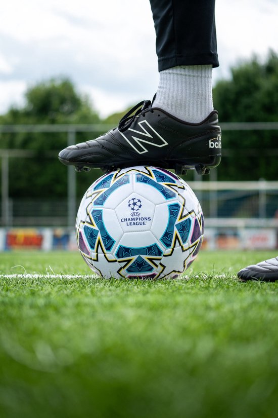 Voetbal met UEFA Champions League logo en sterrenpatroon, met een zwarte voetbalschoen erop. Staand op gras.
