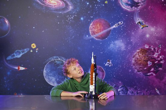 Jongen met krullend haar, kijkt omhoog naar een LEGO Technic NASA Artemis ruimtevaartuig op een zwart platform, met een ruimte-achtergrond.