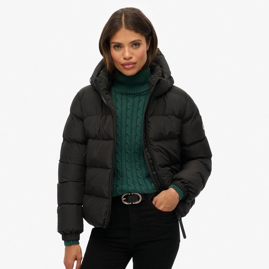Vrouw draagt een zwarte gewatteerde jas met capuchon, een groene coltrui en een zwarte riem.