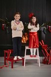 Twee kinderen in kerstsfeer, de jongen draagt een beige kabeltrui en donkerblauwe broek, het meisje een witte trui en rode rok.
