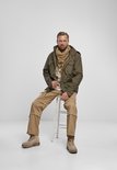 Man zittend in olijfgroene jas, sjaal, gestreept shirt en beige cargo broek.