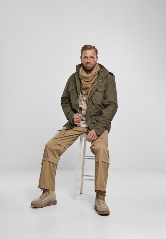 Man zittend in olijfgroene jas, sjaal, gestreept shirt en beige cargo broek.