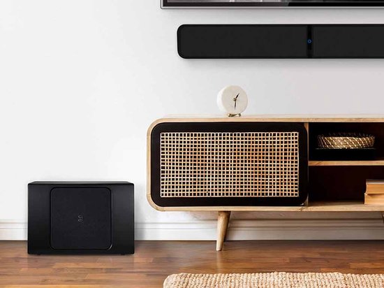 Zwarte Bluesound Pulse Sub+ subwoofer naast een houten tv-meubel in een woonkamer.