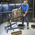 Homme montant un moteur de voiture sur un support moteur dans un atelier, avec des murs à outils.