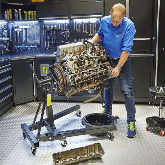 Homme montant un moteur de voiture sur un support moteur dans un atelier, avec des murs à outils.