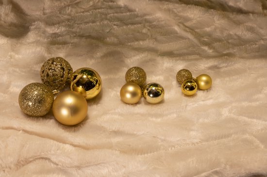 Gouden kerstballen, waaronder glitter en opengewerkte varianten, liggen op een zachte ondergrond.