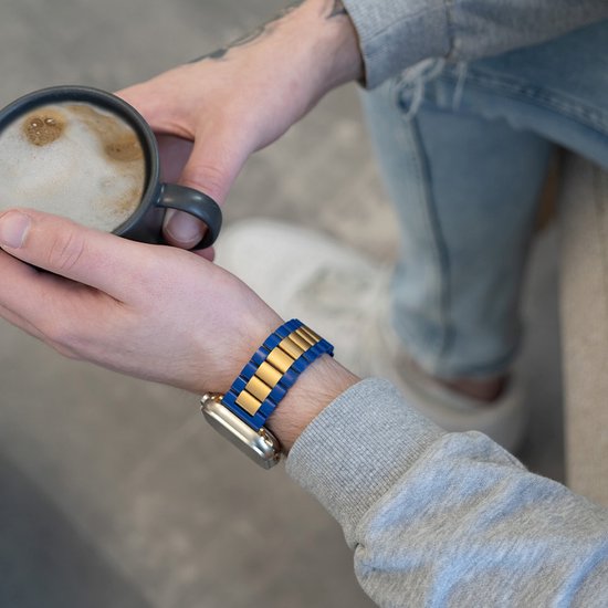 Model draagt een Apple Watch met een blauw en goudkleurig metalen bandje, terwijl hij koffie drinkt.