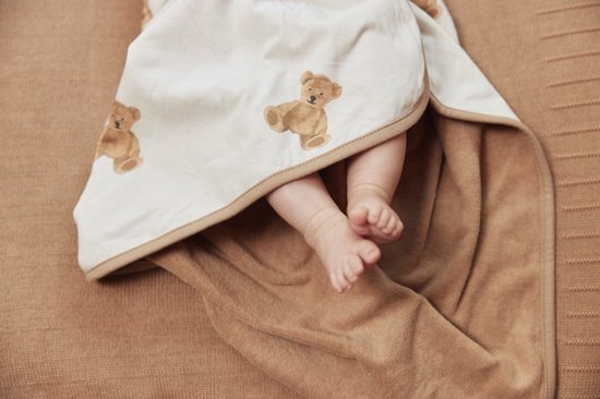 Un bébé aux pieds nus sous une cape de bain marron clair à imprimé oursons. La cape de bain a un bord beige.
