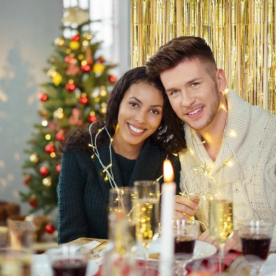 Koppel aan tafel met kerstboom, omringd door lichtslingers.