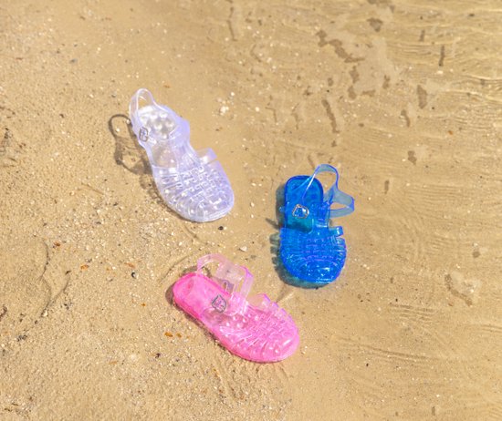 Drie transparante waterschoenen in blauw, roze en paars op zanderig strand.