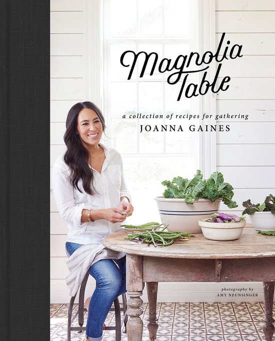 Joanna Gaines zit aan een tafel met een boek "Magnolia Table". Op de tafel liggen groene groenten en paarse aubergines in kommen.