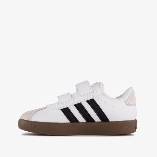 Witte sneaker met zwarte strepen, klittenbandsluiting en bruine zool op witte achtergrond.