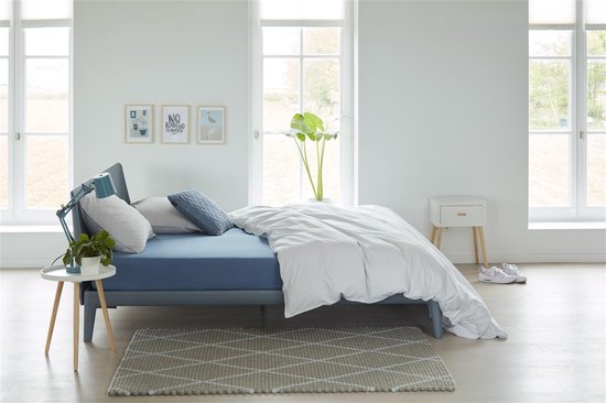 Een opgemaakt bed met blauw hoeslaken, wit dekbed en gestreepte deken in lichte slaapkamer.