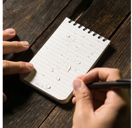 Een hand schrijft met een pen op een Rite in the Rain notebook met waterdruppels erop, liggend op een houten ondergrond.