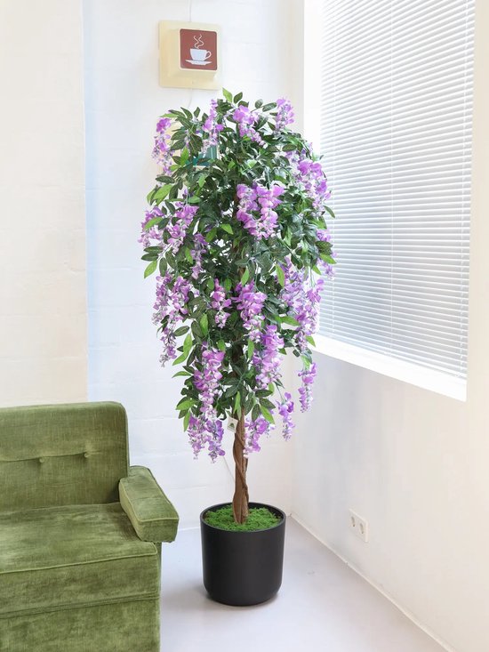 Arbre artificiel de glycine avec fleurs violettes et feuilles vertes dans un pot noir, à côté d'un canapé vert et près d'une fenêtre.