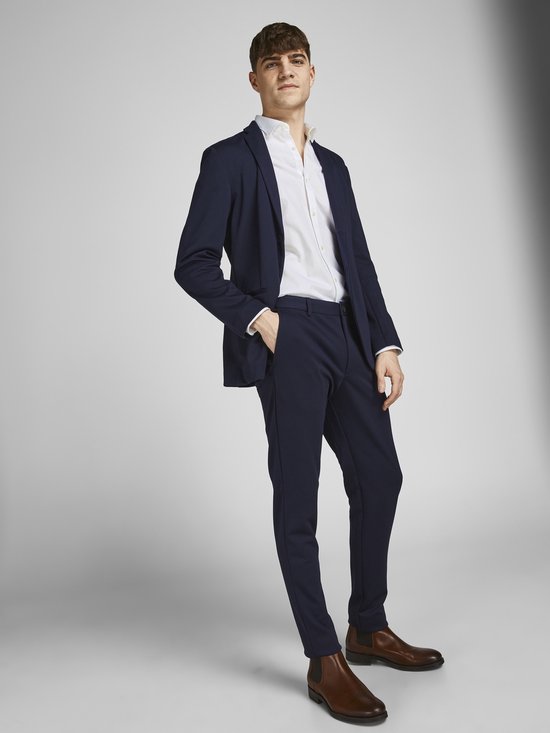 Man met donkerblauwe blazer en broek, wit overhemd en bruine schoenen.