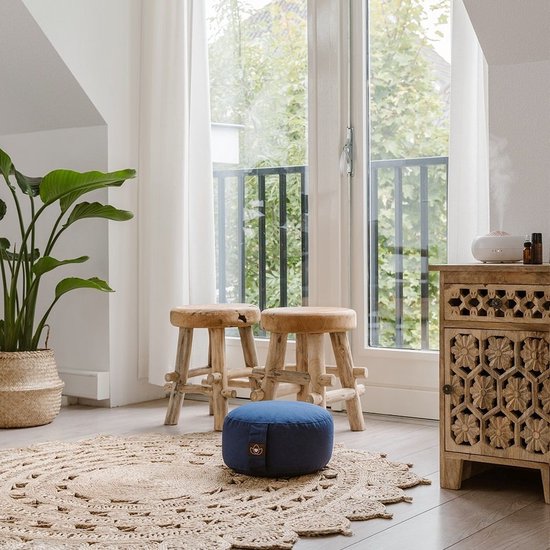 Indigo meditatiekussen op een ronde jute mat in een woonkamer met twee houten krukken en plant.