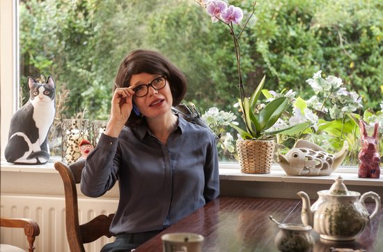 Vrouw met bril in donkerblauw shirt praat aan de telefoon bij een raam met planten en decoraties.