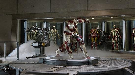 Iron Man in rood en wit pantser op een rond platform in een werkplaats met meerdere identieke pakken op de achtergrond.