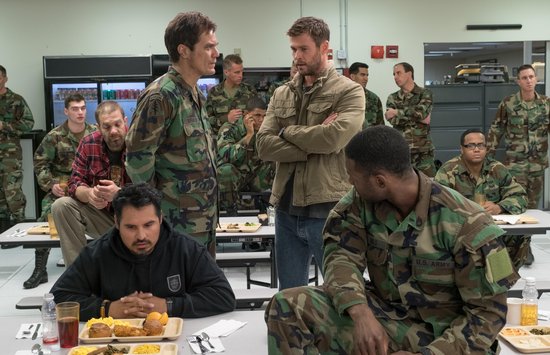Militairen in gevechtskleding zitten aan tafels in een kantine. Chris Hemsworth staat in een bruine jas met armen over elkaar.