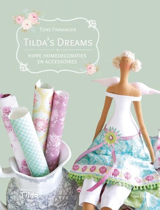 Boek 'Tilda's Dreams' met een Tilda-engel met vleugels, gekleed in een witte jurk en een vrolijke, turquoise en roze rok, zittend naast een theepot vol met opgerolde stof in bloemenpatronen.