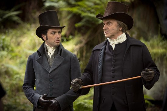 Twee mannen in historische kleding met hoge hoeden en donkere jassen staan buiten in de natuur.