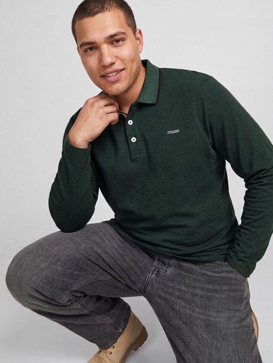 Man van voren gezien, die in de camera kijkt, in een donkergroene polo met lange mouwen en grijze jeans.